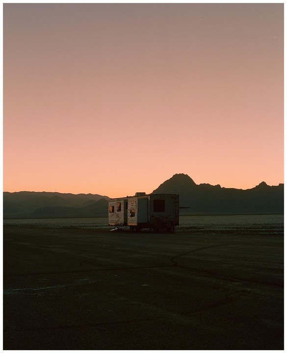 A camper trailer is parked in a vast, flat landscape with mountains in the background under a pink sky.