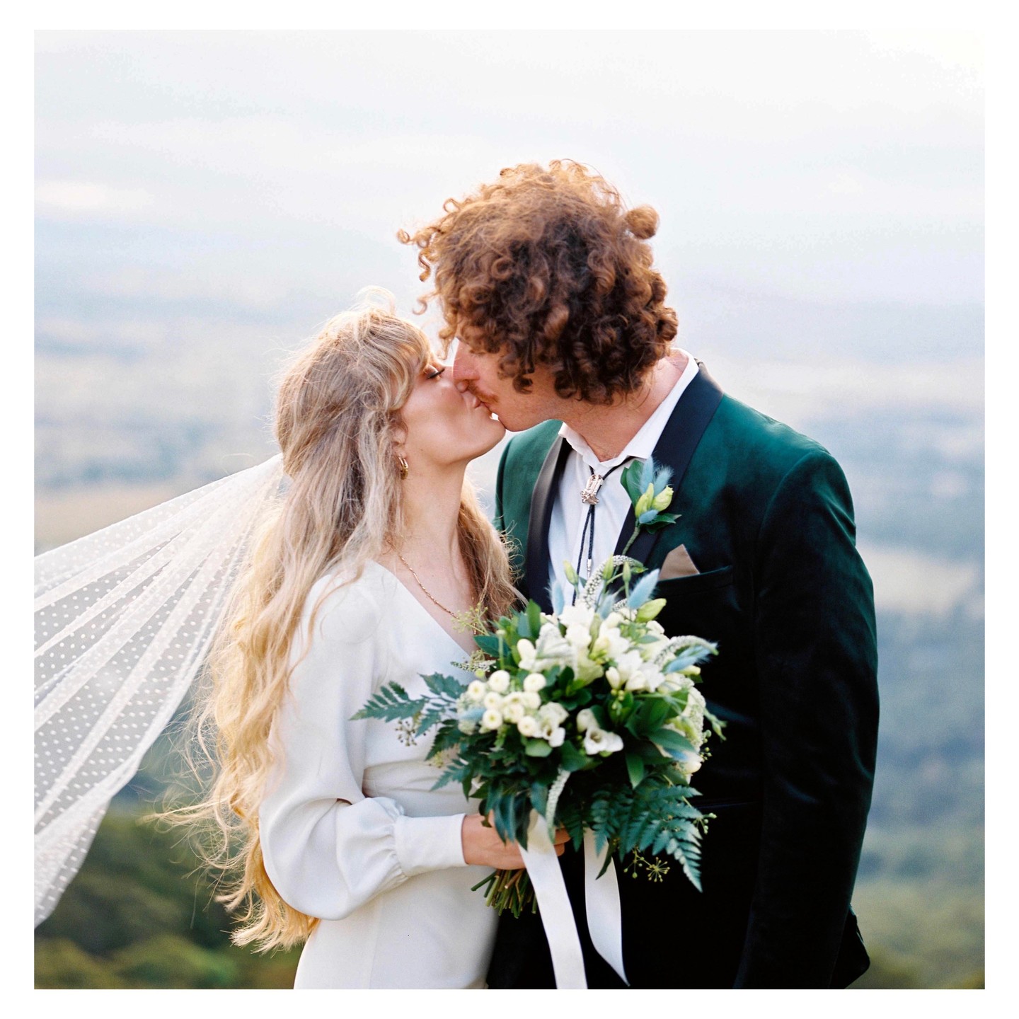 A couple kisses while the woman holds a bouquet of white flowers and green foliage.