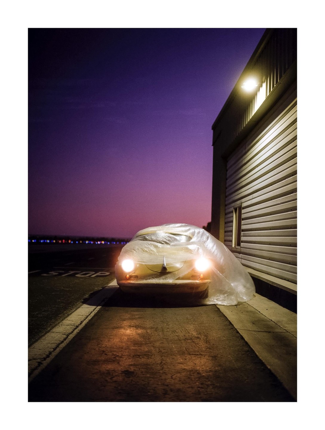 A car covered in plastic sheeting with its headlights on is parked next to a building under a purple sky at night.
