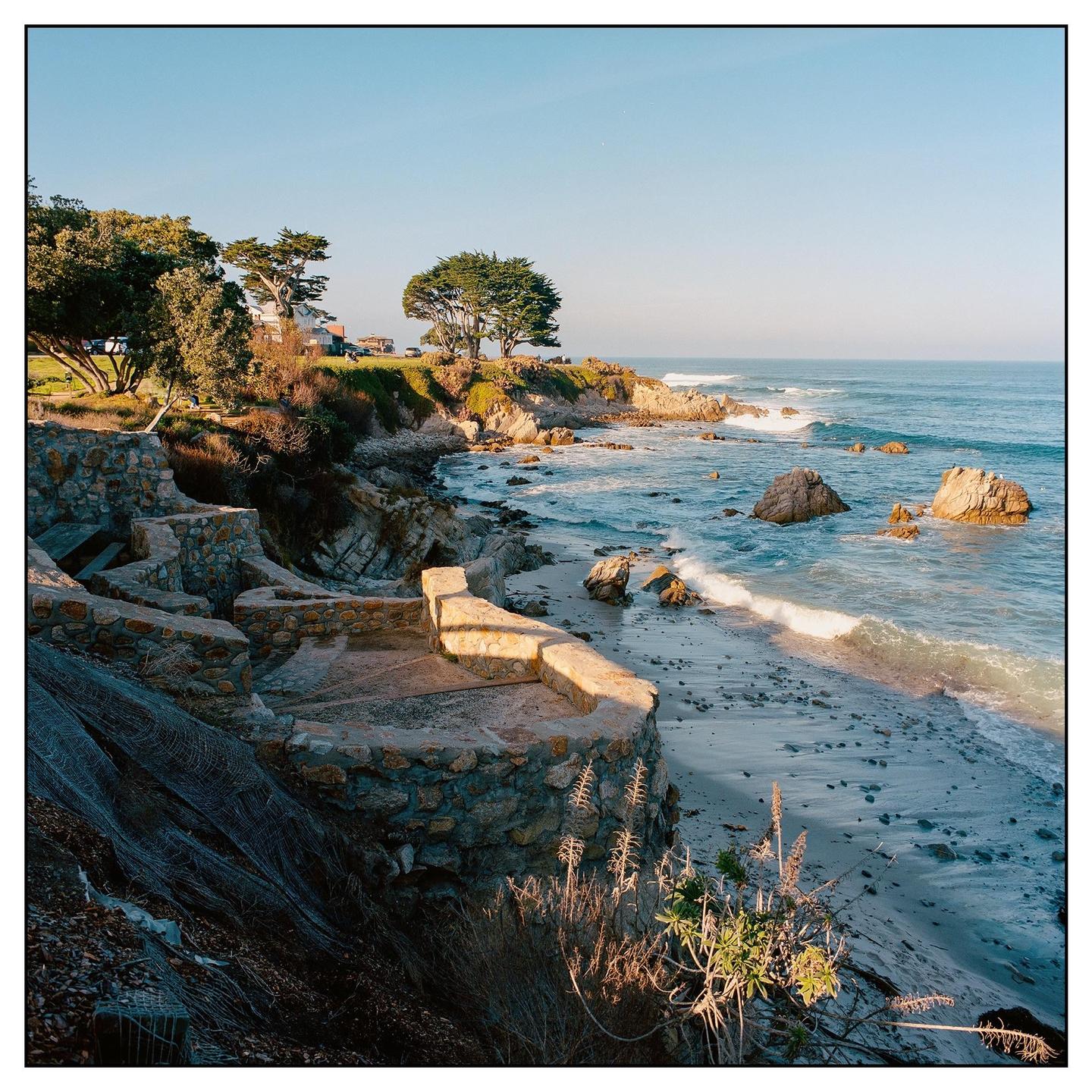 A stone wall and steps lead down to a sandy beach with waves crashing on the shore.