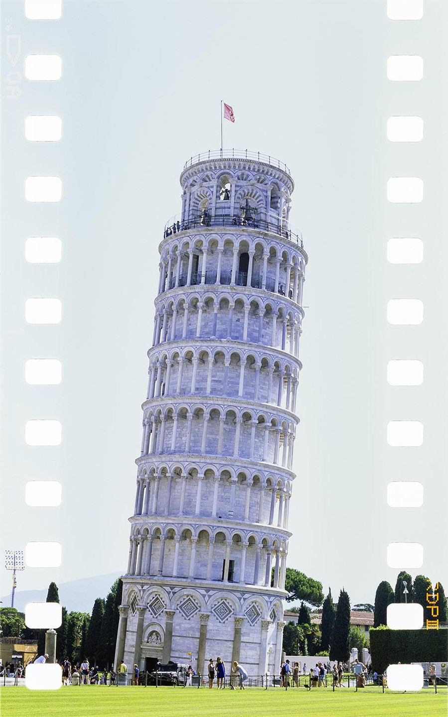 The Leaning Tower of Pisa stands tall with a red flag on top, surrounded by a green lawn and people.