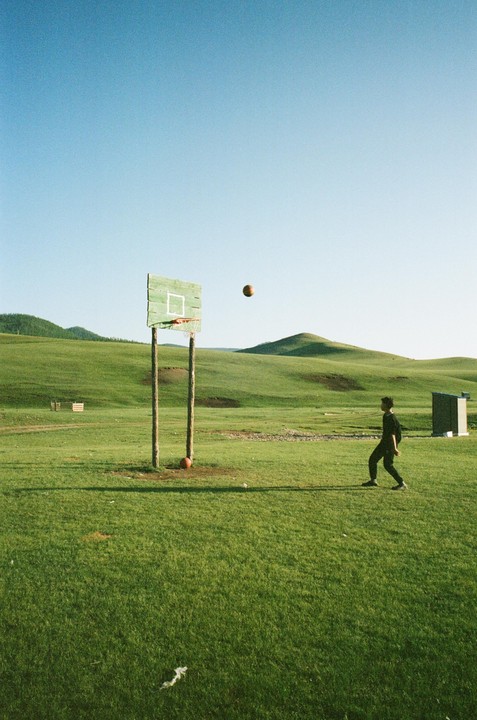 A boy plays basketball on a grassy field with rolling hills in the background.