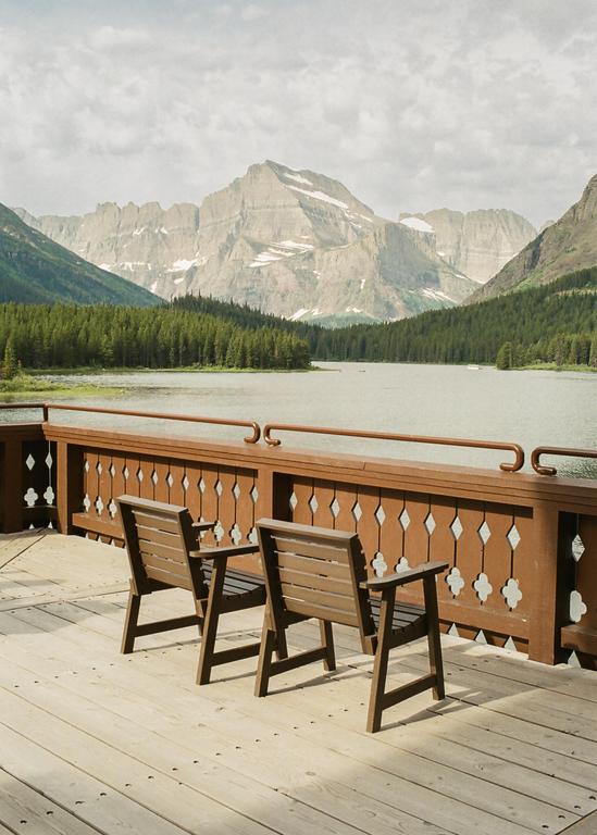 Two wooden chairs sit on a deck overlooking a lake surrounded by a forest and mountains.