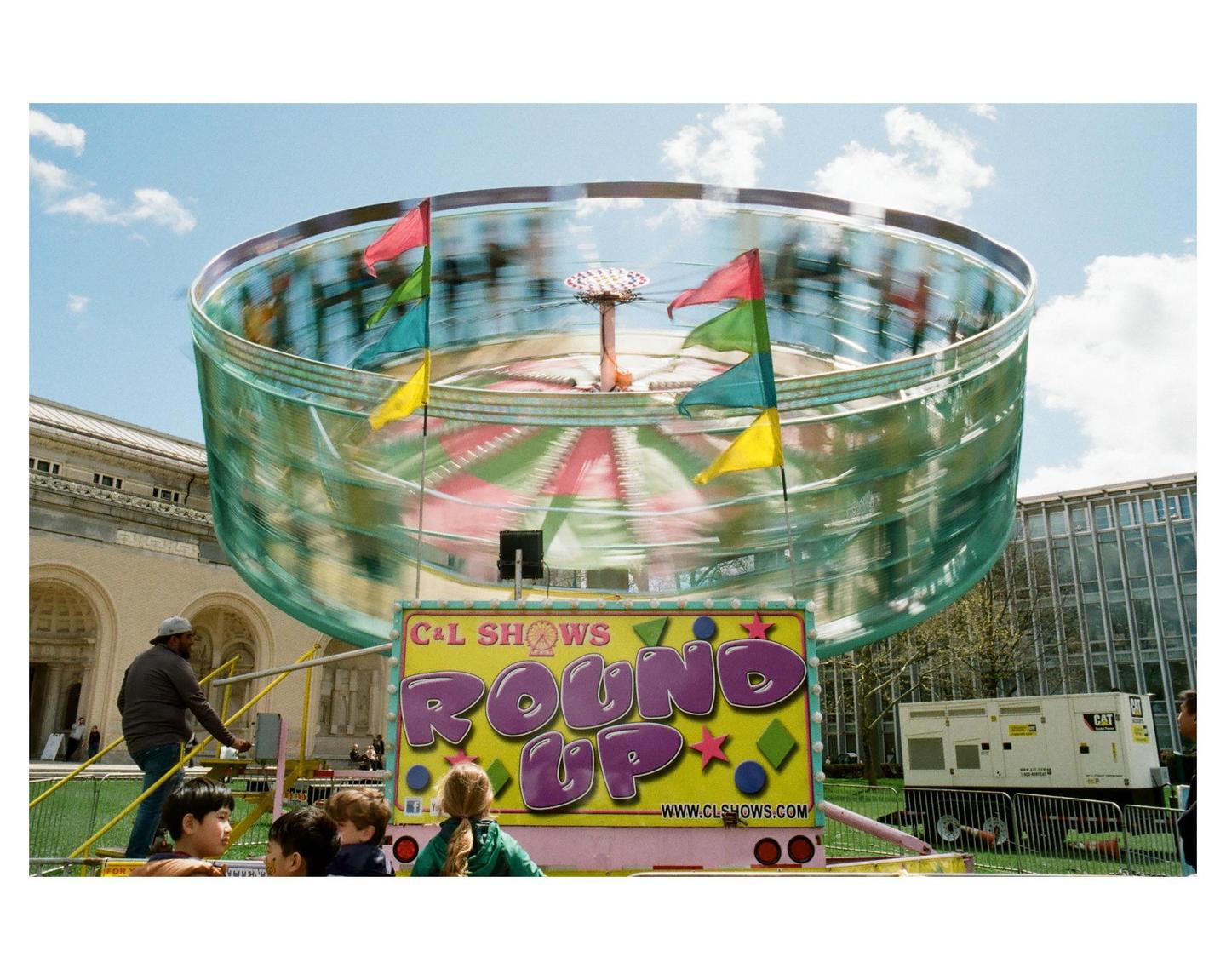 A man operates a spinning carnival ride with children watching in the foreground.