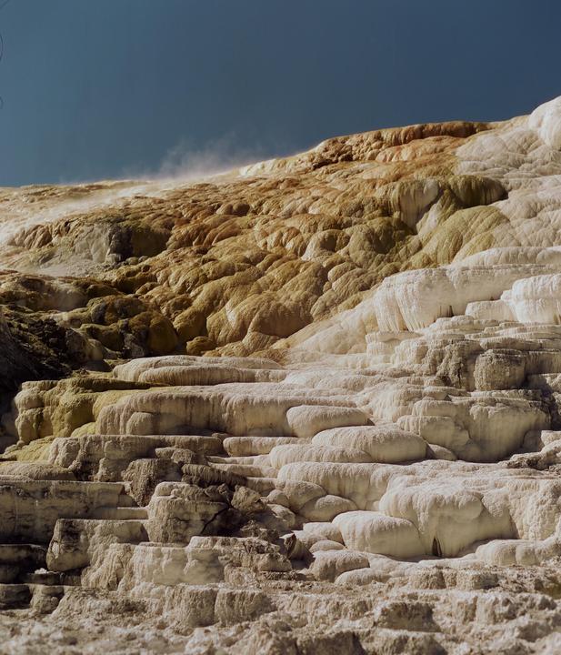 A large, terraced rock formation with white and brown mineral deposits has steam rising from its upper slopes.