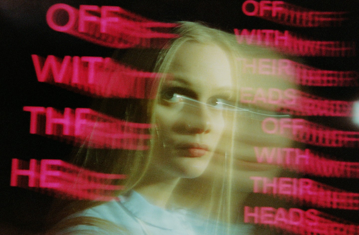 A woman with long blonde hair looks up and to the right, with red text that reads "OFF WITH THEIR HEADS" blurred in the background.