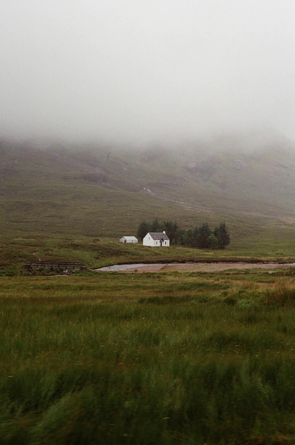 A small white house and shed sit in a green valley with a river and bridge, shrouded in fog.