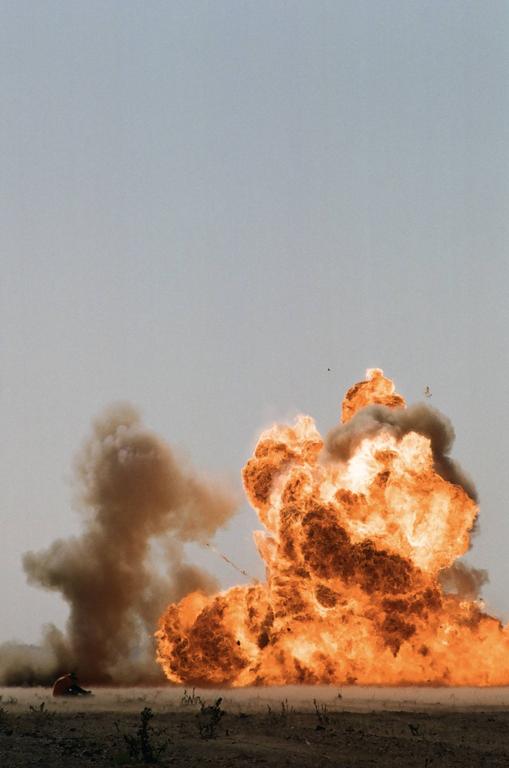 A large explosion with fire and smoke rises from the ground, with a person in the distance.