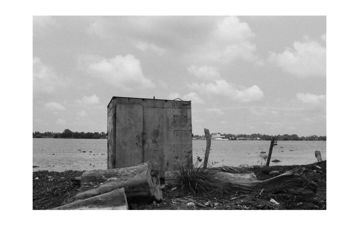 A small, weathered shack stands on the shore of a body of water, with trees and buildings visible in the distance under a cloudy sky.