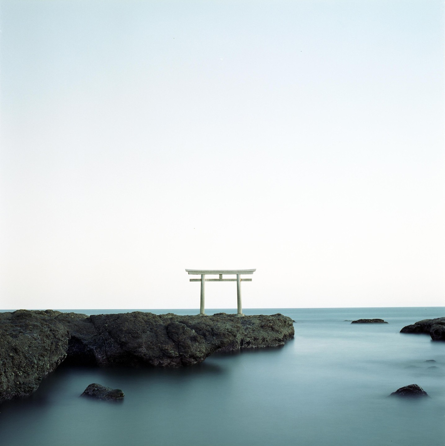 A torii gate stands on a rocky outcrop in the ocean under a bright sky.