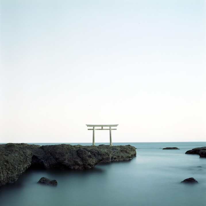 A torii gate stands on a rocky outcrop in the ocean under a bright sky.