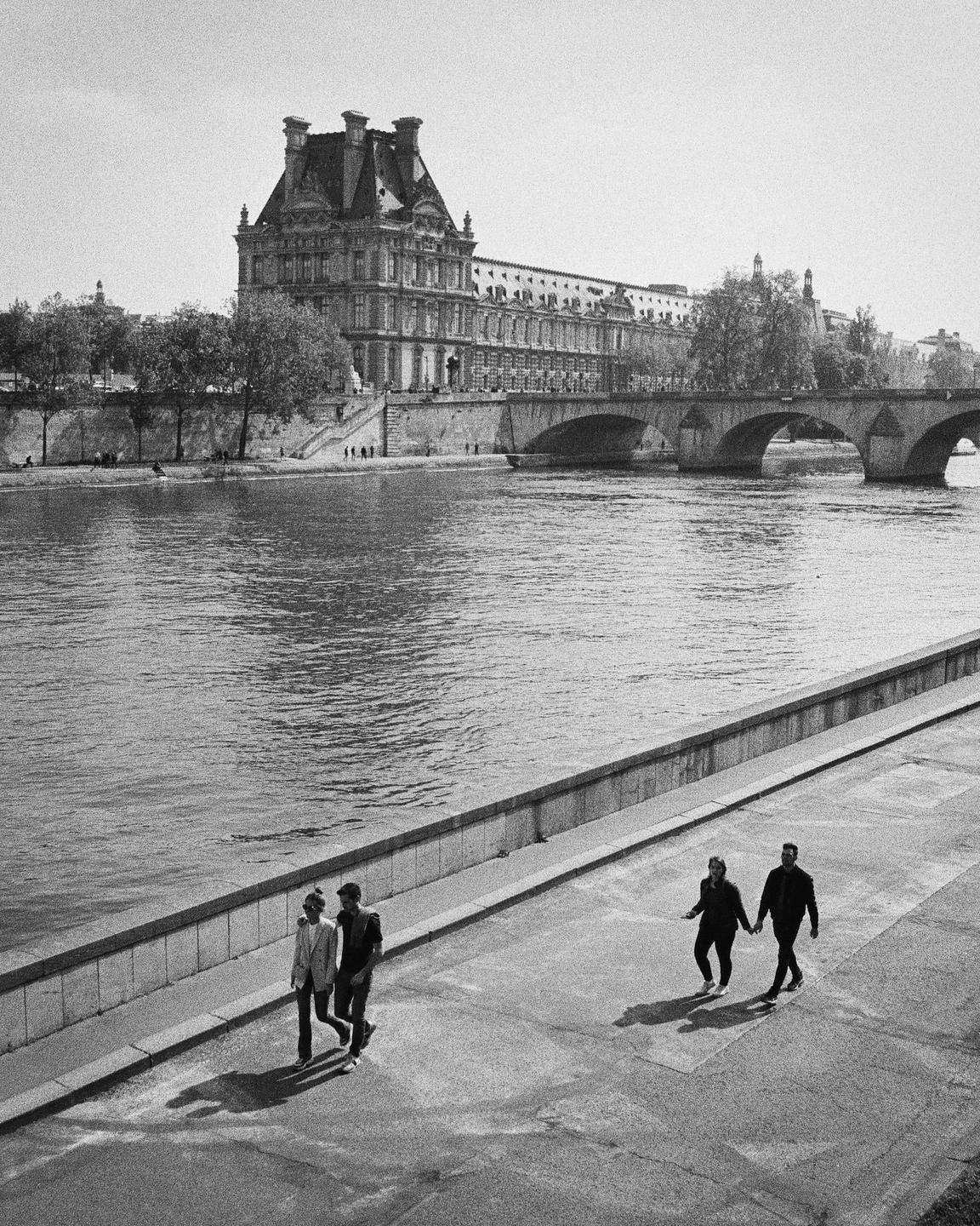 Two couples walk along a river with a large building and bridge in the background.