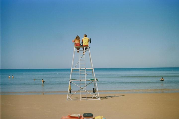 A couple sits on a lifeguard stand overlooking a beach with people swimming in the ocean.
