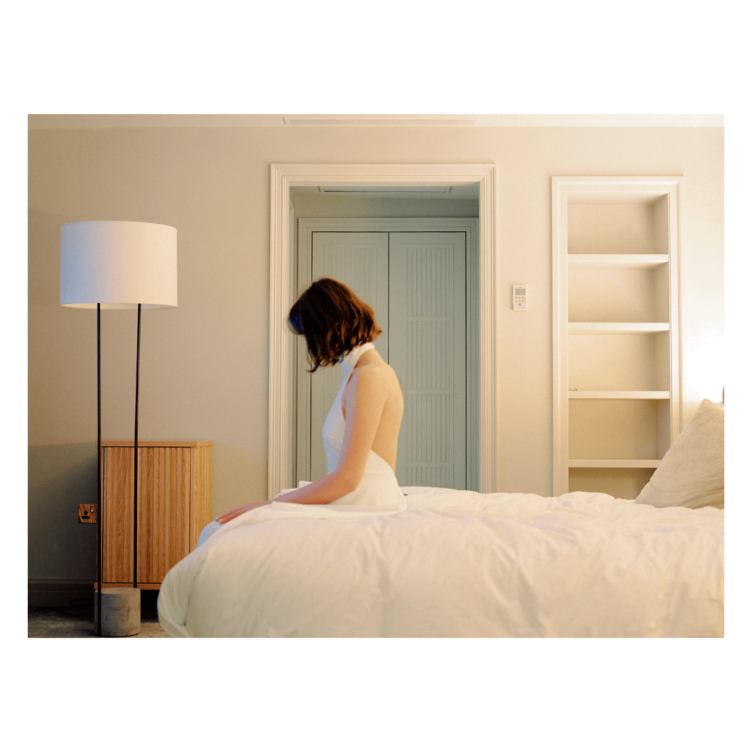 A woman in a white dress sits on a bed in a bedroom with a lamp and a bookshelf.