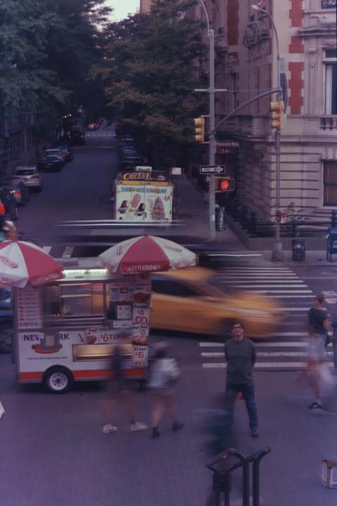 A man stands in the middle of a crosswalk as blurred people and a taxi pass by, with food carts and buildings lining the street.
