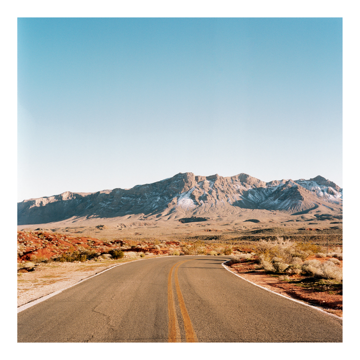 A winding road leads towards a mountain range with patches of snow under a clear sky.