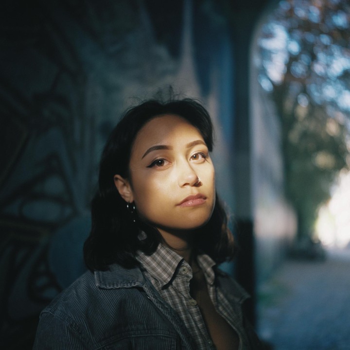 A woman with dark hair and a nose ring looks directly at the camera, with a beam of light illuminating her face against a dark, graffiti-covered wall.