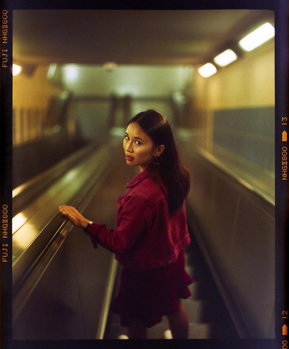 A woman in a red jacket and skirt looks back while riding an escalator in a dimly lit tunnel.