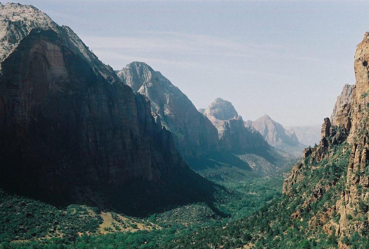 A valley with green trees and bushes is surrounded by tall, rocky mountains under a hazy sky.