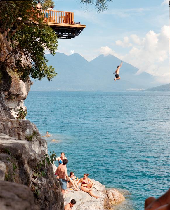 A man jumps from a wooden platform into a large body of water with mountains in the background while other people relax on rocks by the shore.