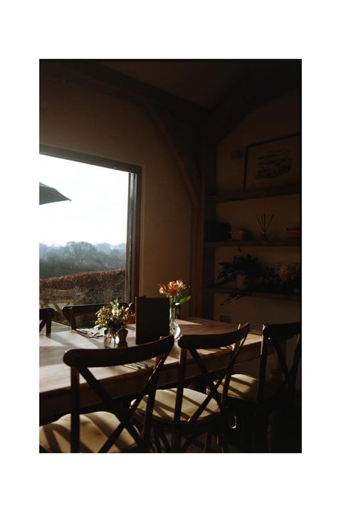 A dining table with chairs and flower arrangements sits next to a window overlooking a hazy, tree-covered landscape.
