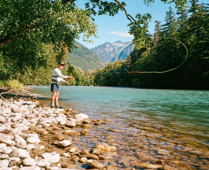 A man fly fishes in a river with mountains and trees in the background.