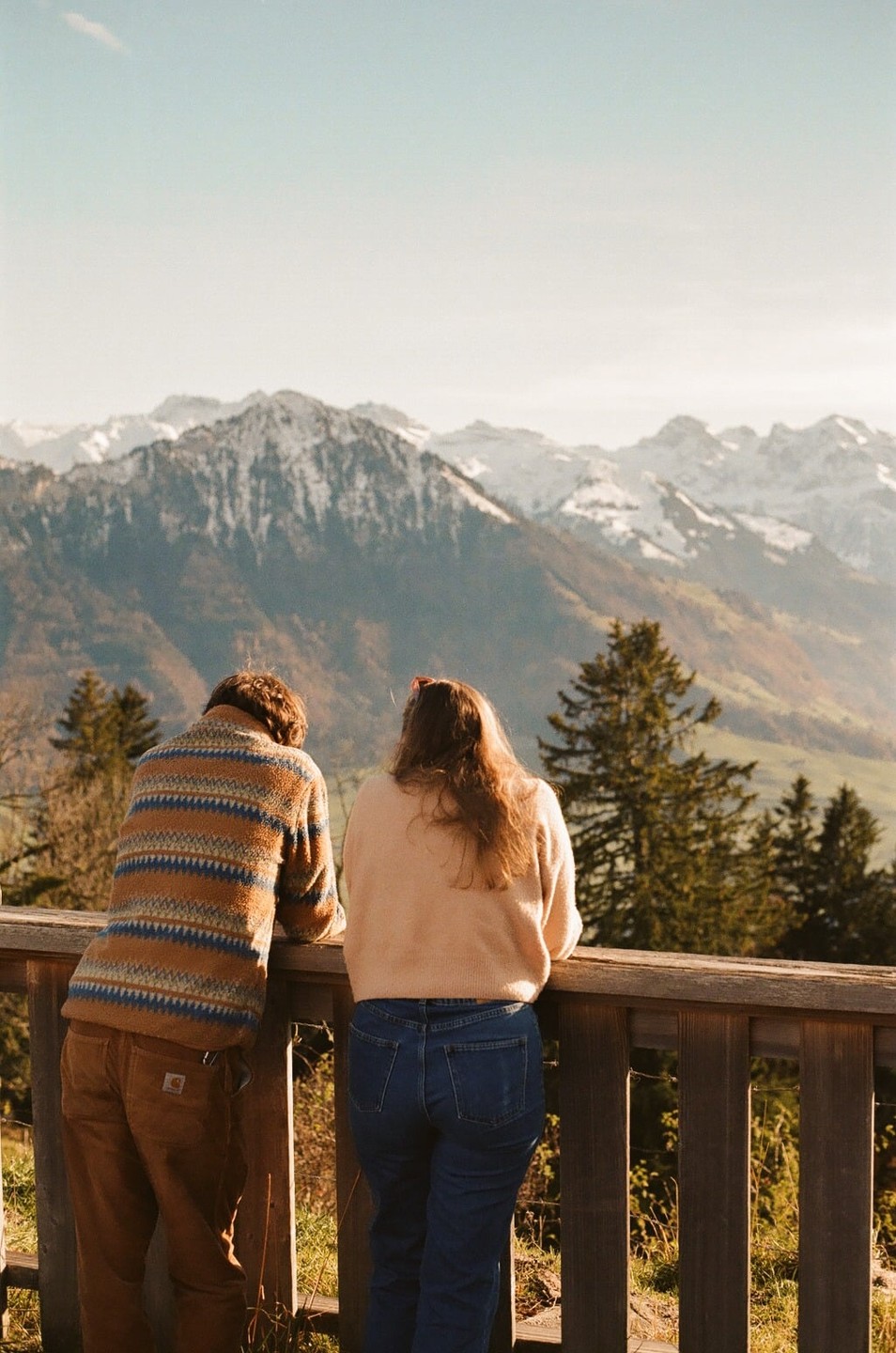 A couple stands on a wooden balcony overlooking a valley with snow-capped mountains in the distance.