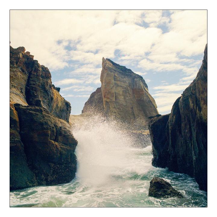 A wave crashes against a rocky cliff face, with a large rock formation in the background under a cloudy sky.