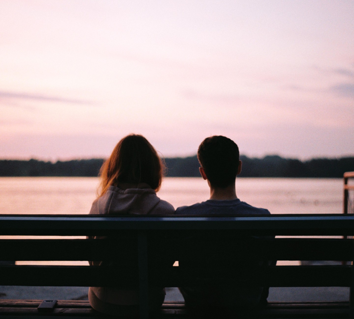 A couple sits on a bench looking out at a body of water with a pink sky at dusk.