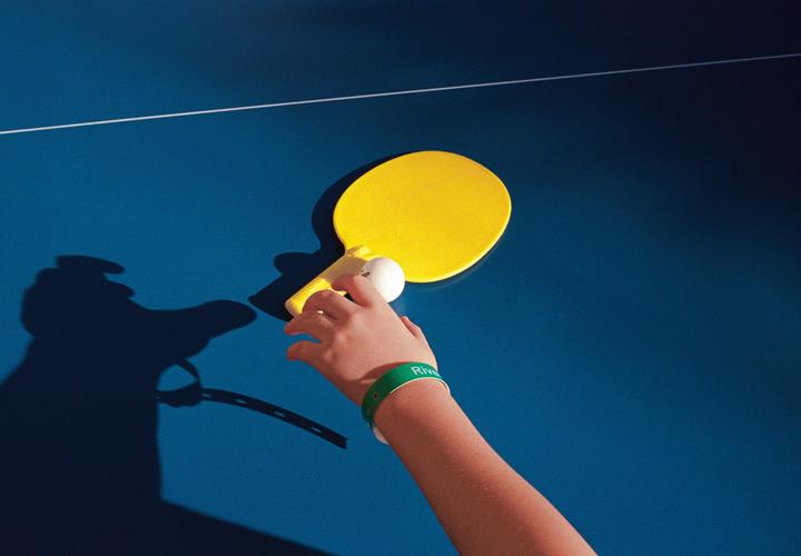 A child's arm with a green wristband holds a yellow ping pong paddle and a white ball over a blue table.