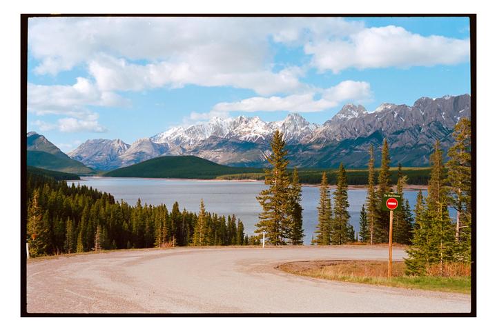 A winding road overlooks a lake surrounded by a forest with snow-capped mountains in the distance under a blue sky with clouds.