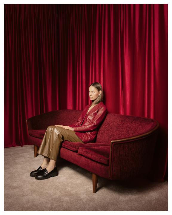 A woman in a red leather jacket and brown pants sits on a red velvet couch in front of a red curtain.