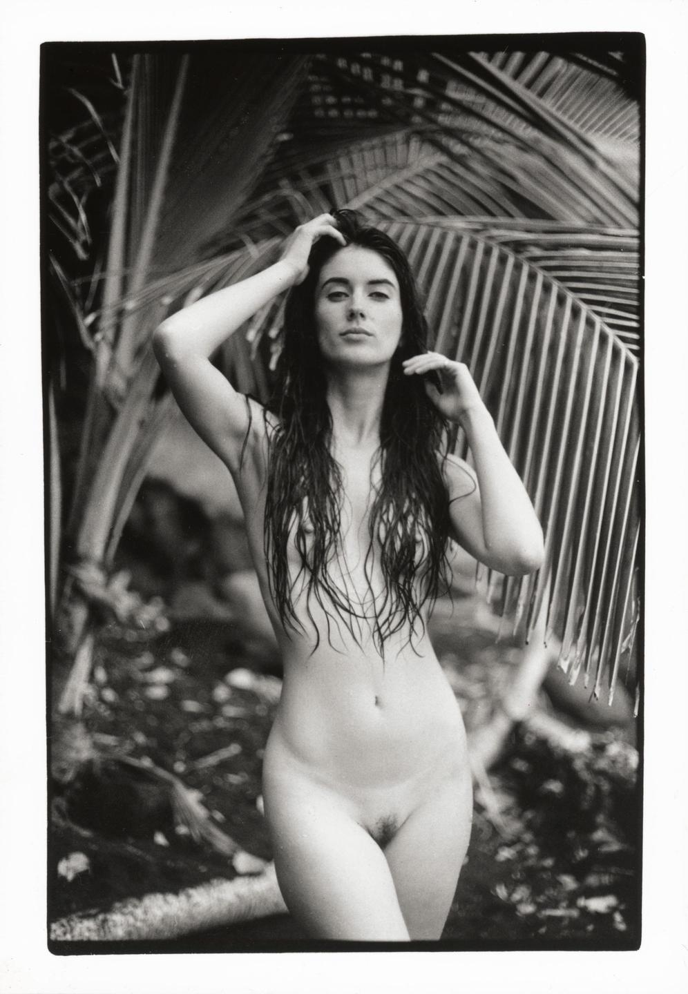 A nude woman with long wet hair stands in front of palm fronds.