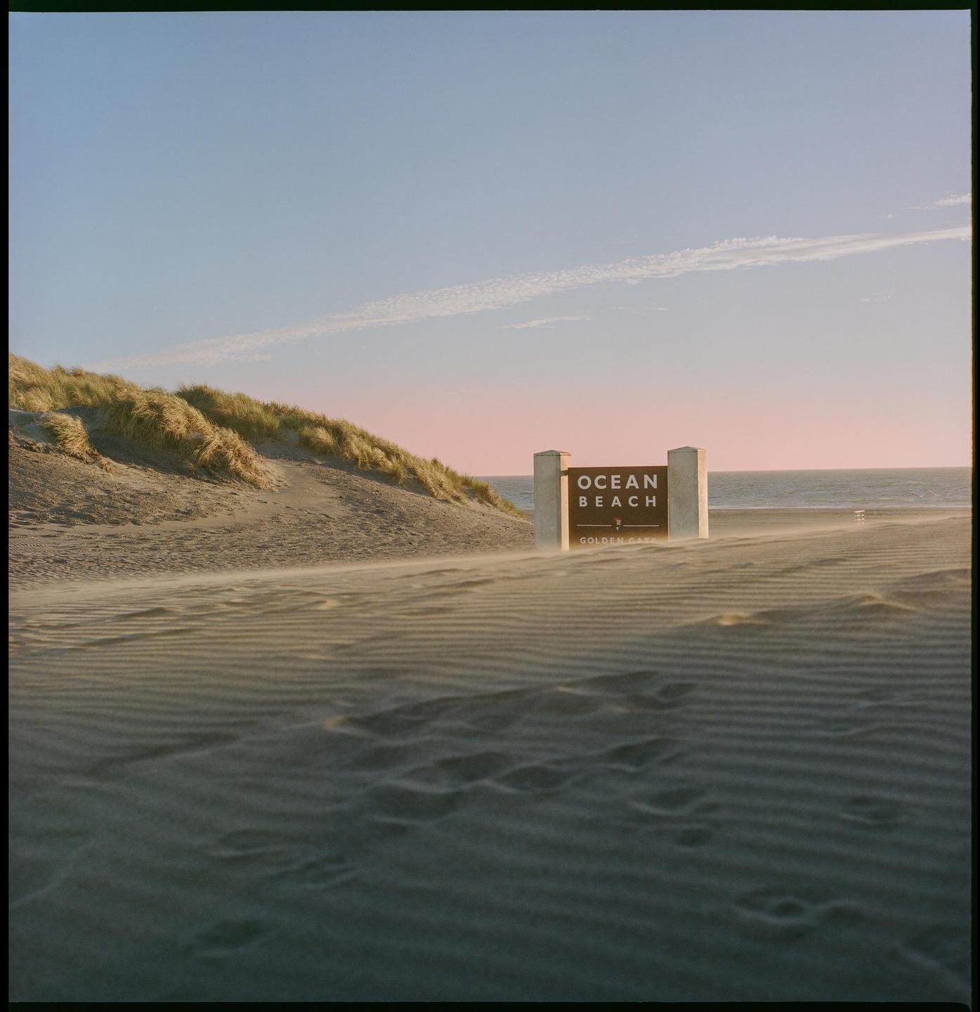 A sign for Ocean Beach stands on a sandy beach with dunes and the ocean in the background.