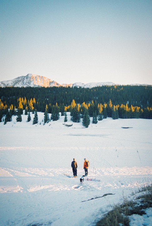 Two men and a dog stand on a snow-covered field with skis, facing a forest and mountains in the distance.