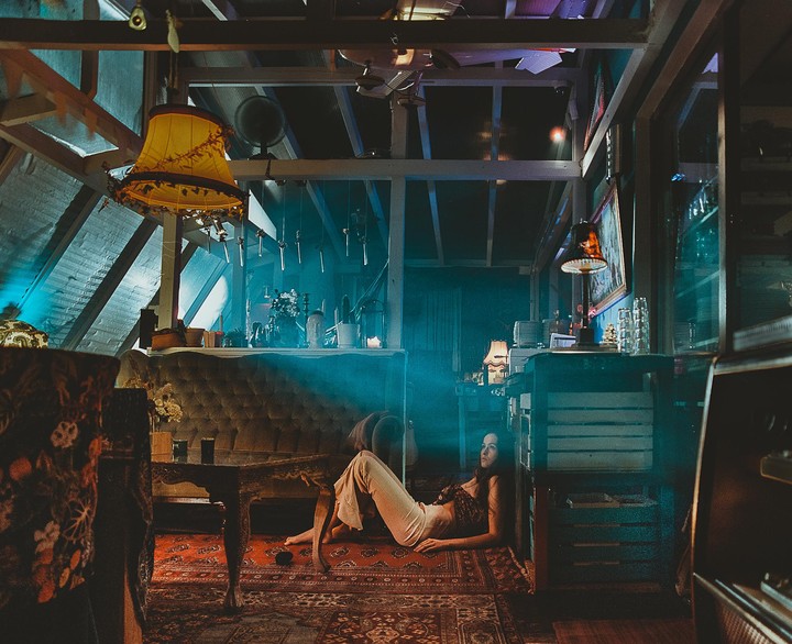 A woman reclines on a patterned rug in a dimly lit room with exposed wooden beams and various lamps.