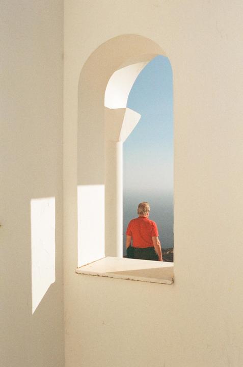 A man in a red shirt looks out through an arched window at a blue sky and distant water.