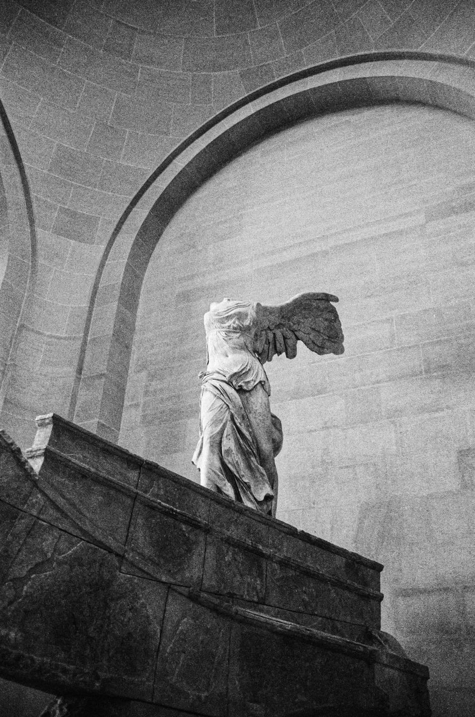 A headless winged statue stands on a pedestal in a large room with arched ceilings.