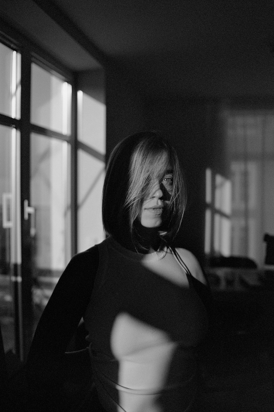 A woman with short hair stands in a room with sunlight casting shadows across her face and body.