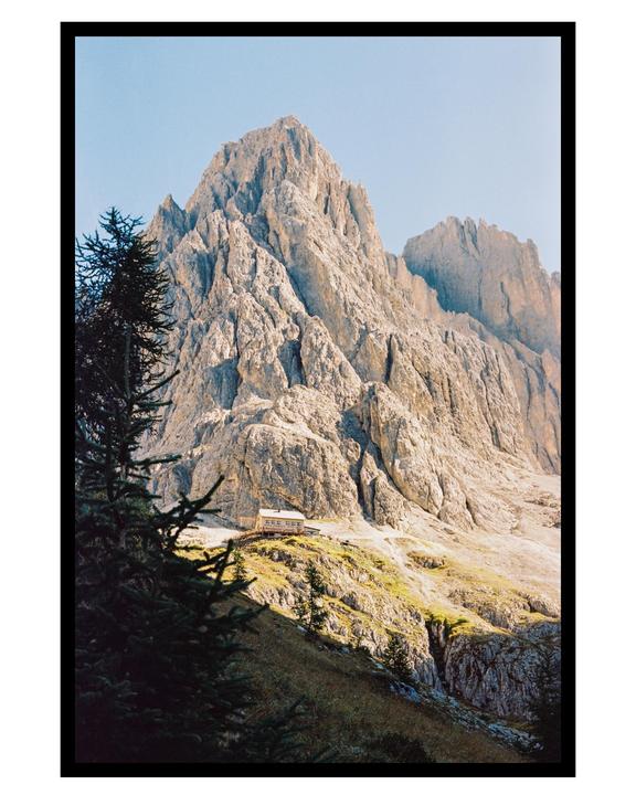 A small building sits on a grassy slope at the base of a large, rocky mountain.