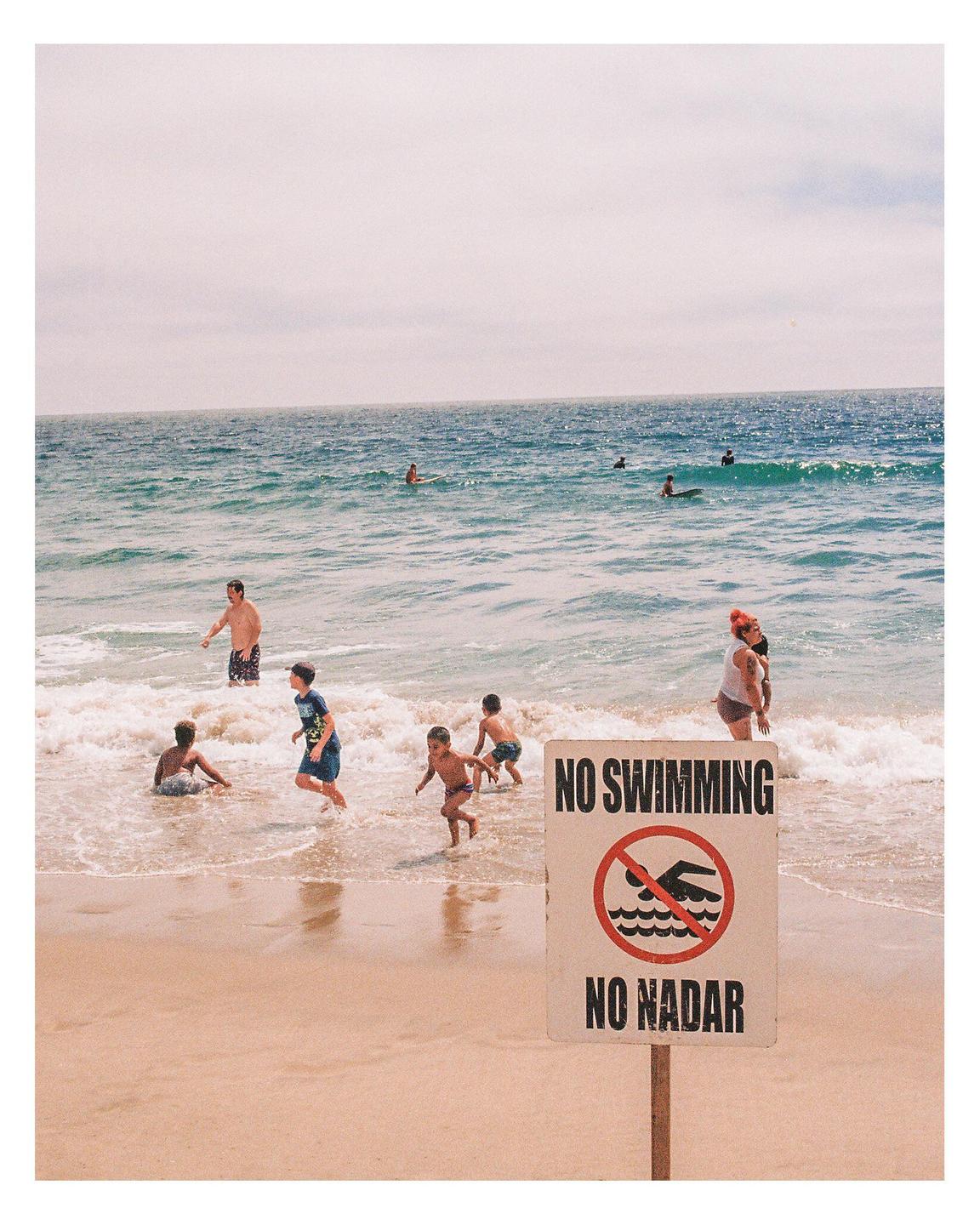 A "No Swimming" sign stands on a sandy beach with people in the ocean.