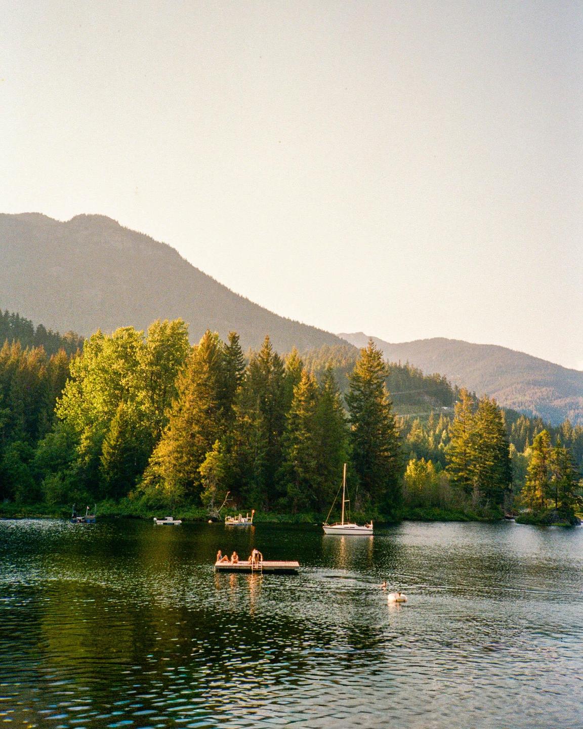 A lake with boats and a floating dock with people is surrounded by a forest and mountains.