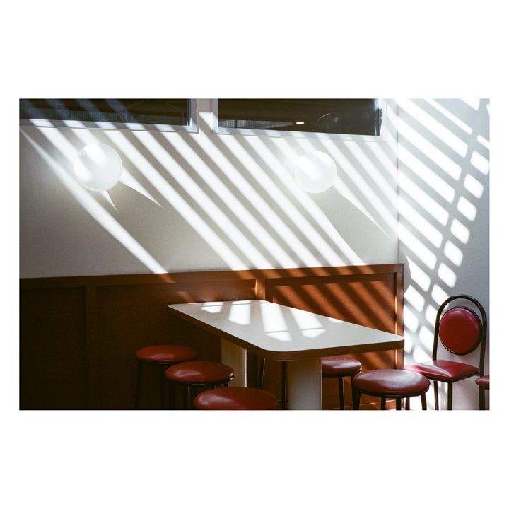 Sunlight streams through a window, casting striped shadows across a diner booth with a table and red stools.