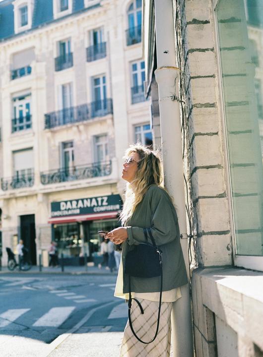 A woman with long wavy hair and sunglasses stands on a sunny street next to a building, holding a phone and a handbag.