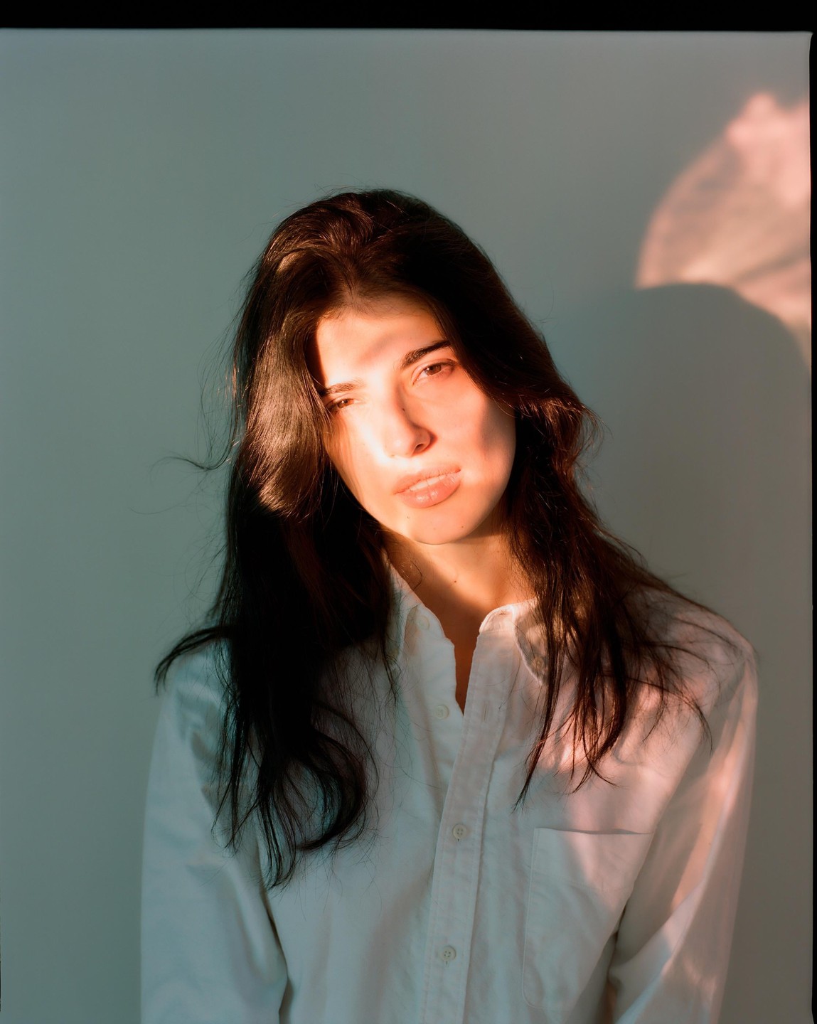 A woman with long dark hair wears a white shirt, with a pool of light on her face and shoulder.