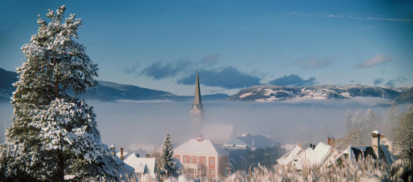 A snow-covered town with a prominent church steeple is nestled in a valley filled with fog, surrounded by snow-capped mountains under a blue sky.