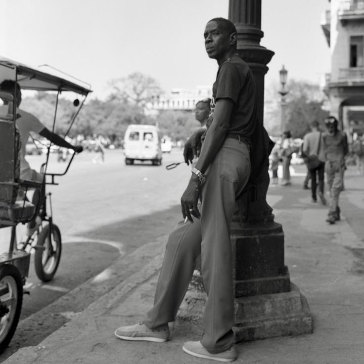 A man leans against a lamppost on a street in Havana, Cuba, with a cigarette between his fingers.