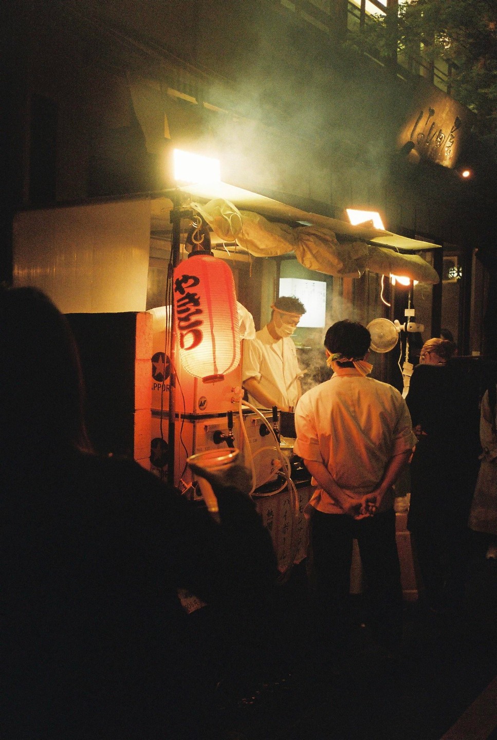 A street food vendor prepares food at night under a red lantern and bright lights, with people watching.