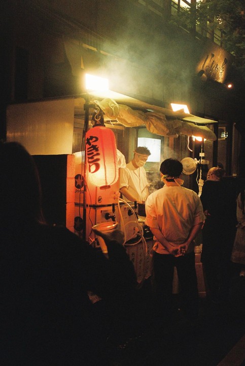 A street food vendor prepares food at night under a red lantern and bright lights, with people watching.
