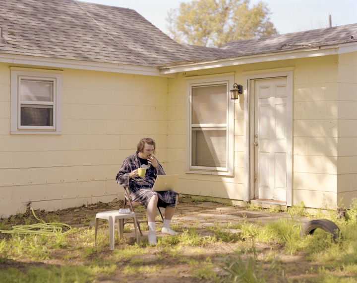 A man in a robe sits in a yard with a laptop, smoking and drinking from a mug.
